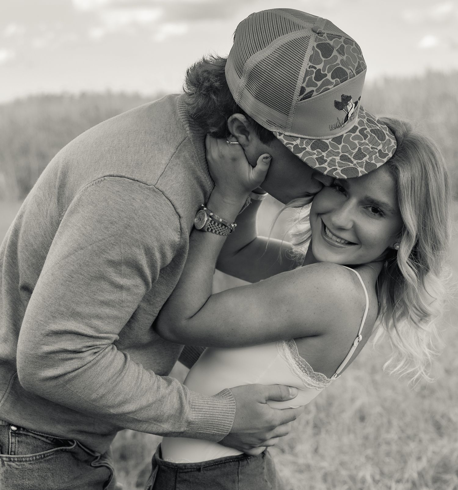 Black and white photo of a couple embracing outdoors, man in trucker hat kissing womans cheek as she smiles.