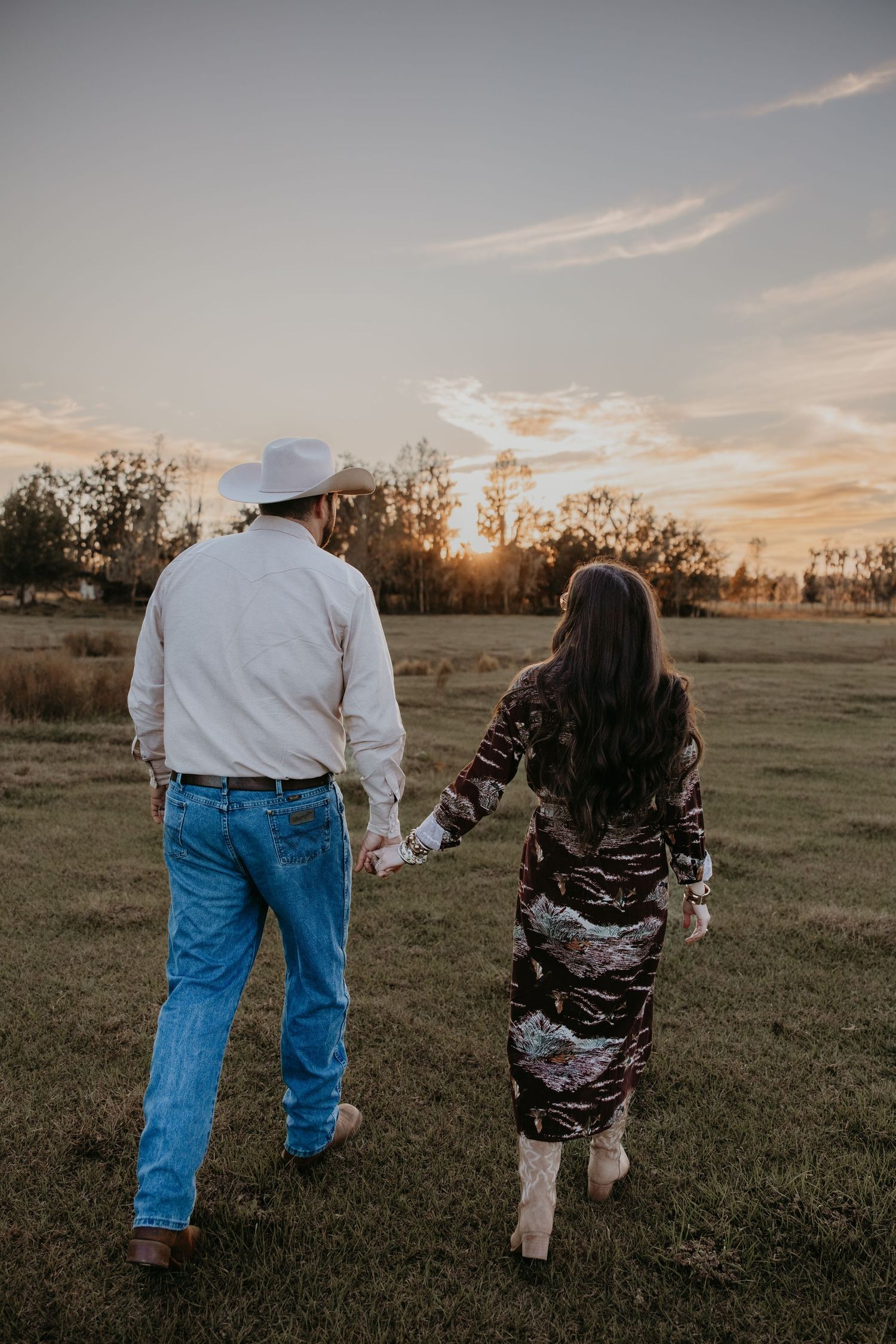 Couple holding hands walking through a green field at sunset, man in white cowboy hat and jeans, woman in floral dress.