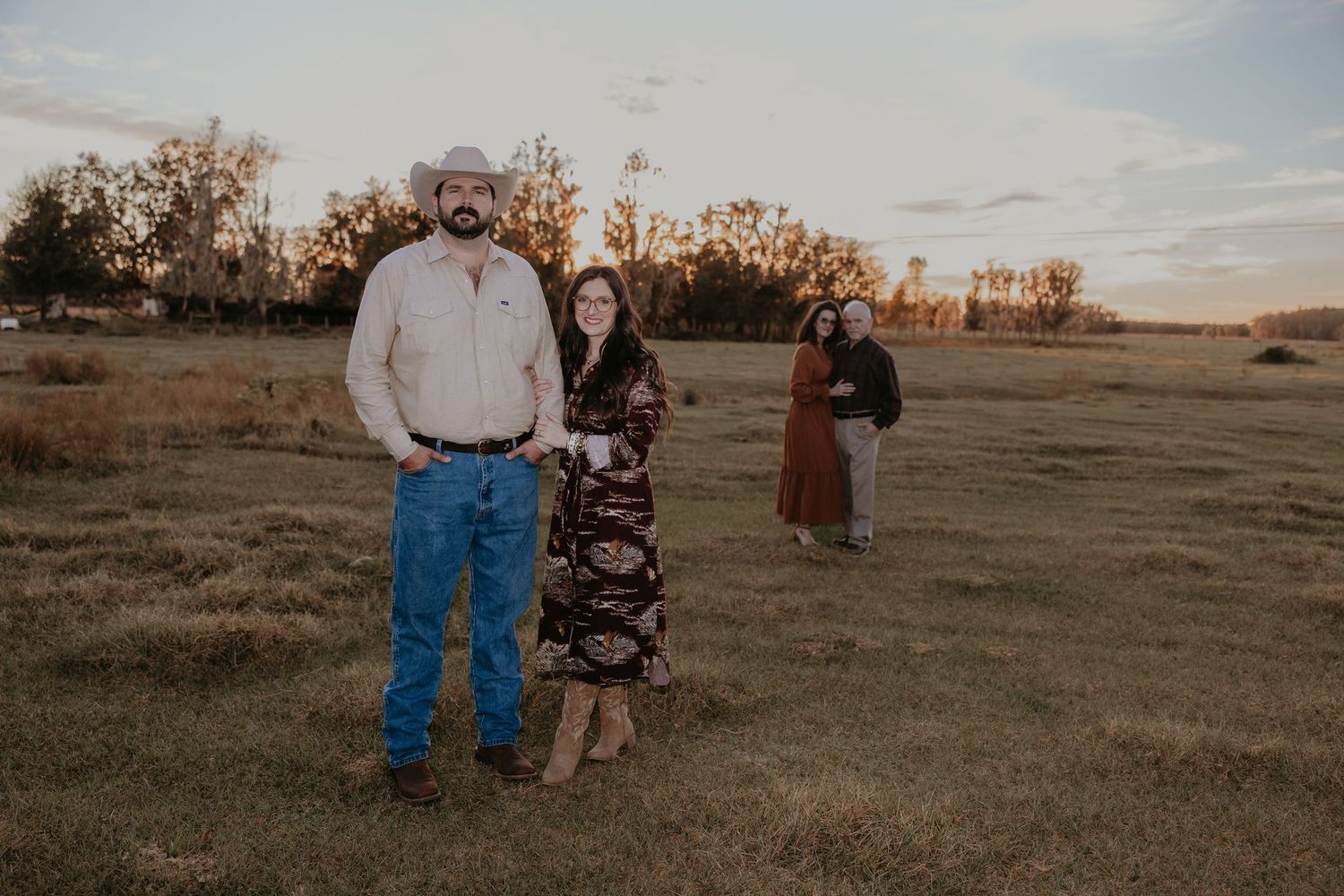 Couple embracing in golden field at sunset, woman in denim skirt and boots, man in cap and jeans.