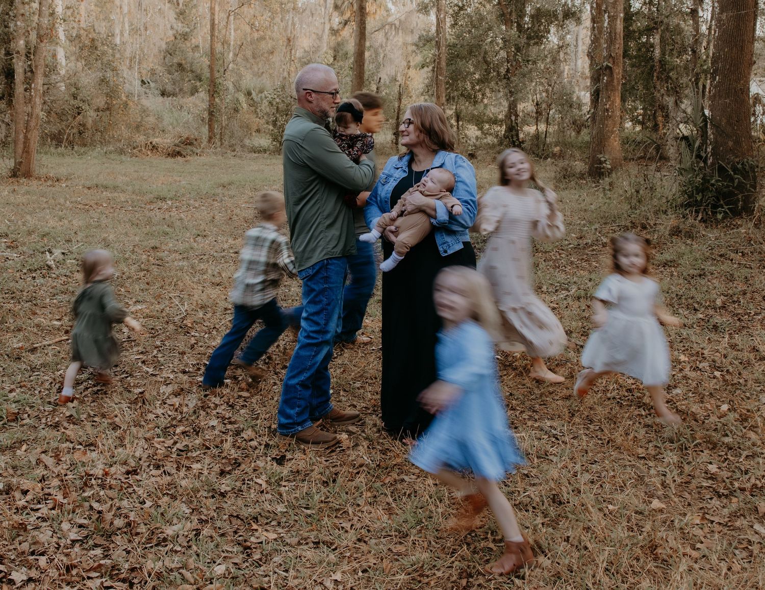 Family photo session in autumn woods with parents holding baby while children play around them.