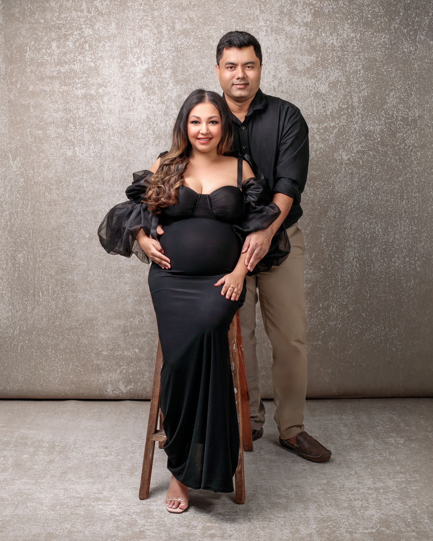 Mum sits on a ladder and partner embraces her from behind in the studio, mum wearing a modern black dress, fine art maternity photo.