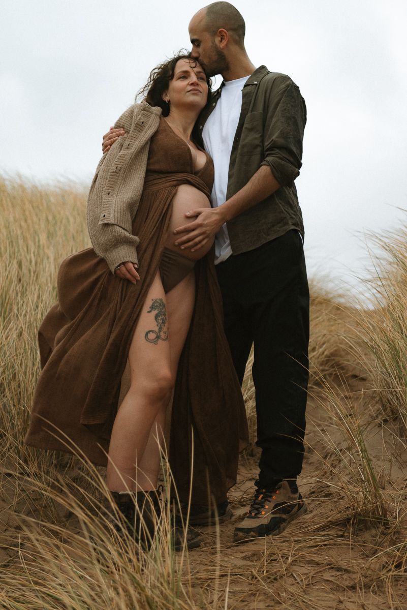 A romantic maternity photoshoot in a field of tall grass shows an intimate moment between a couple at sunset.