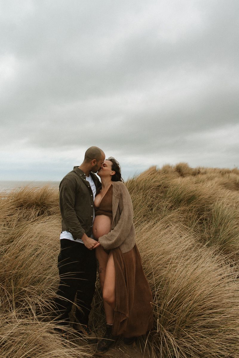 A romantic maternity photoshoot in tall grass on a moody overcast day with swaying beige grass fields.