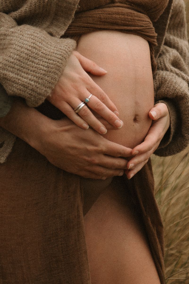 Close-up of hands cradling pregnant belly with engagement ring visible, wearing brown knit sweater.