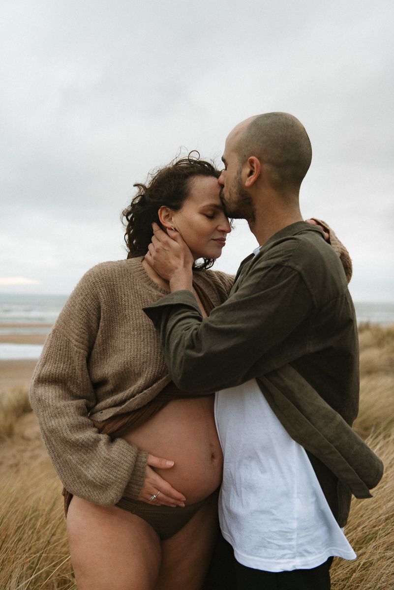 A tender maternity moment shared between a couple embracing on a windswept beach with tall grasses.