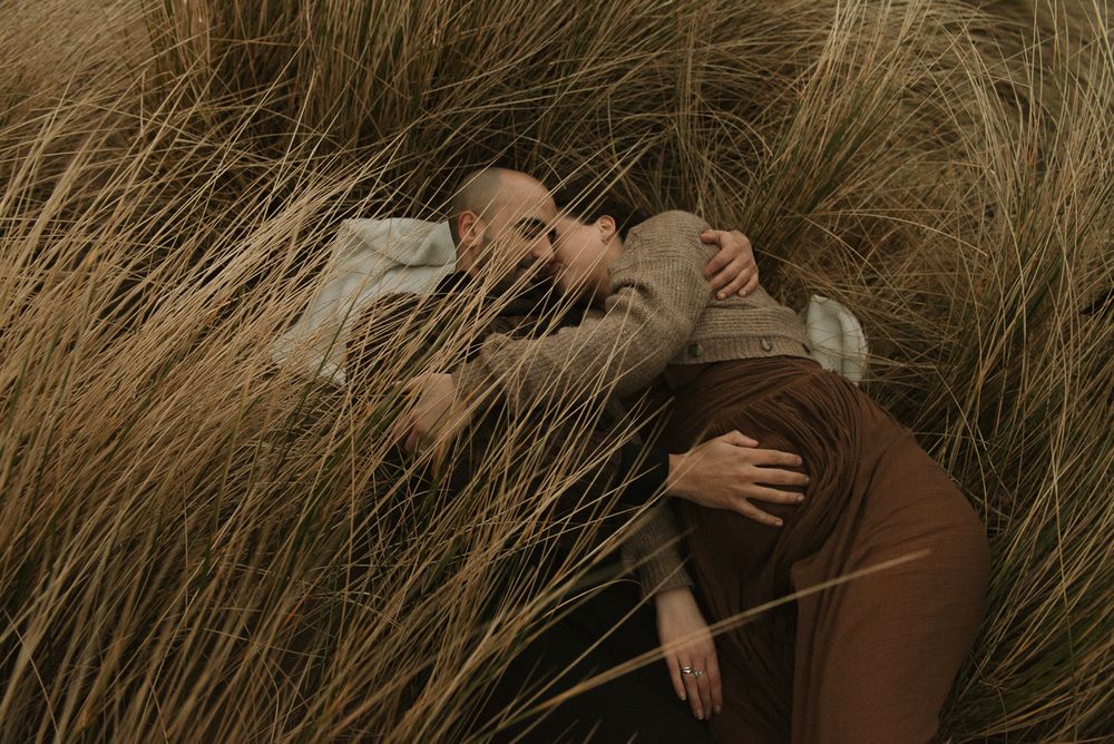 Two people embracing while lying in tall brown autumn grass during a moody outdoor photoshoot.