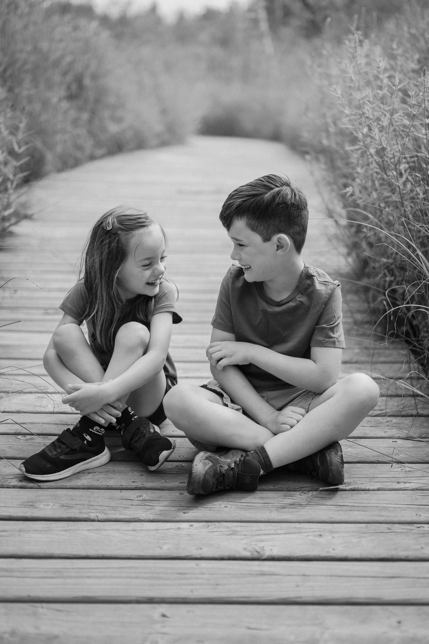 Two children sit cross-legged on a wooden boardwalk sharing a laugh together.