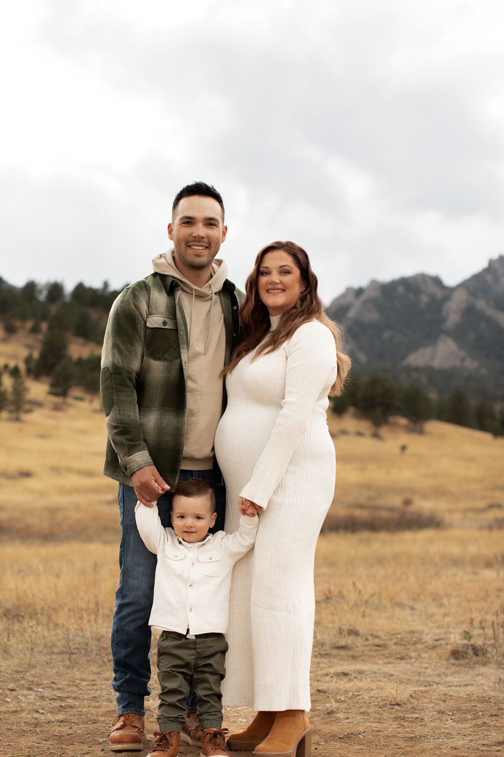 Family of three poses in a grassy field with mountains in the background during maternity photoshoot.