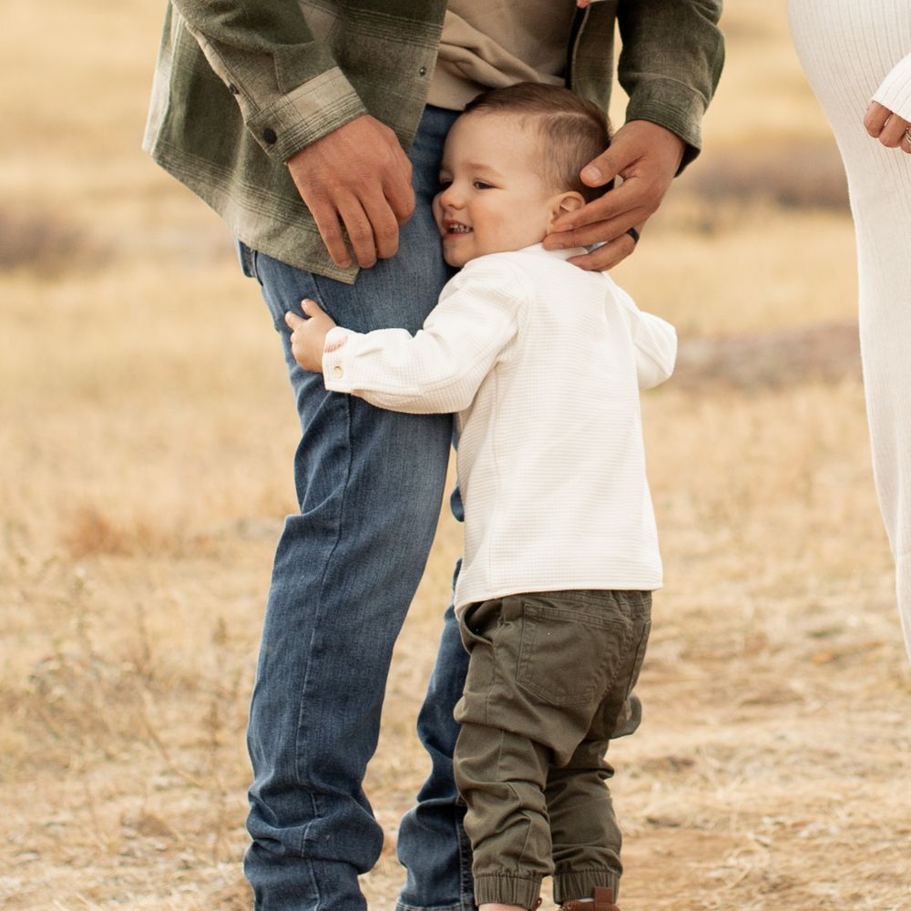 A toddler in a white sweater hugs an adult's legs while standing in a golden field during sunset.