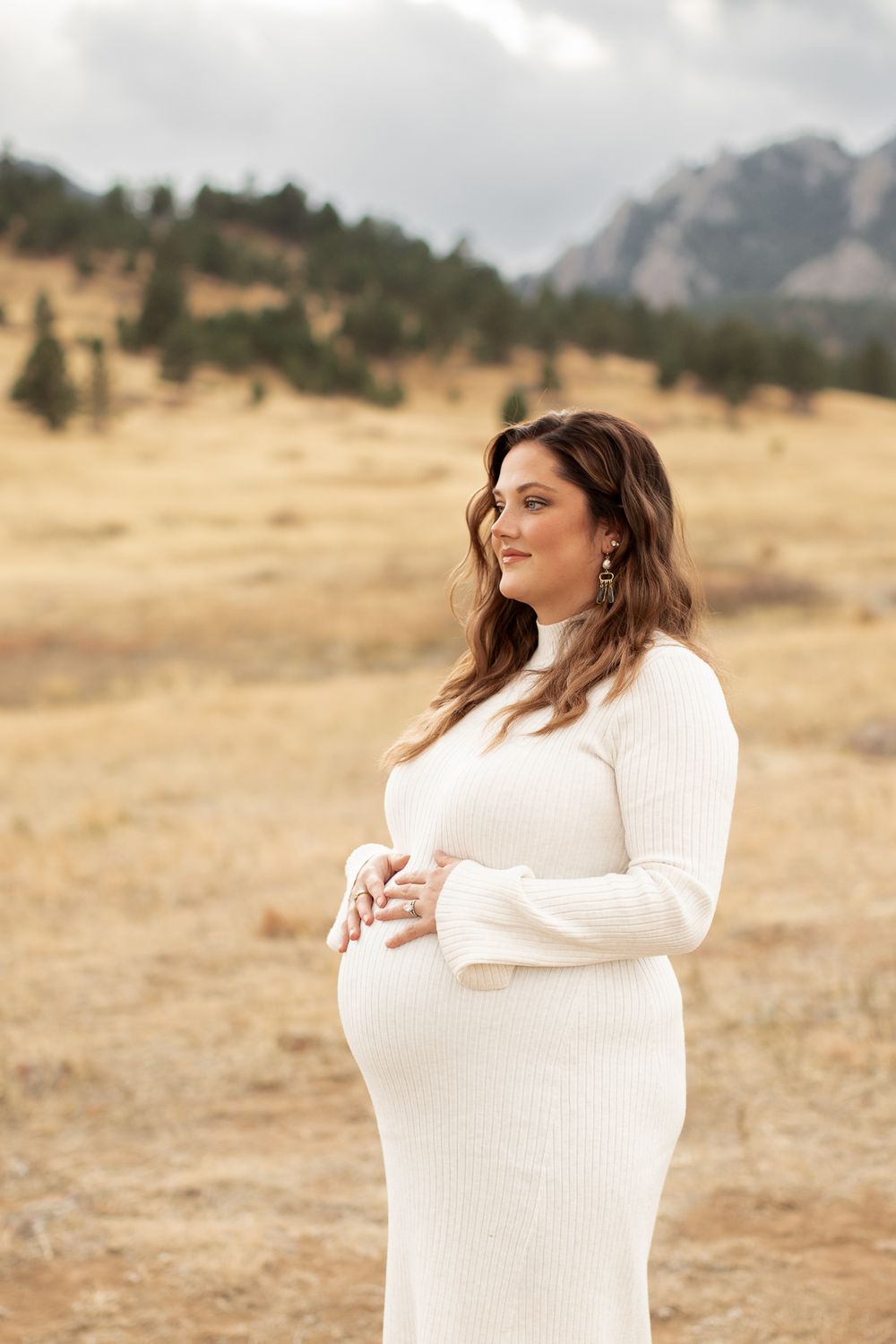 Expectant woman in white sweater dress stands in golden field with mountains in background during overcast day.
