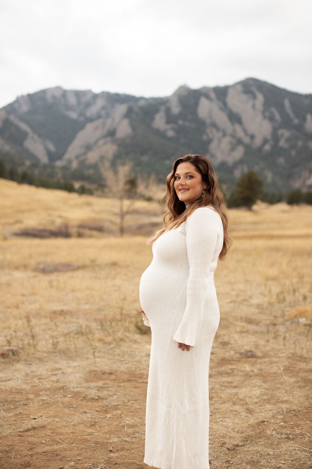 A figure in a white long dress stands in golden fields with the Flatirons mountain range of Boulder, Colorado in background.