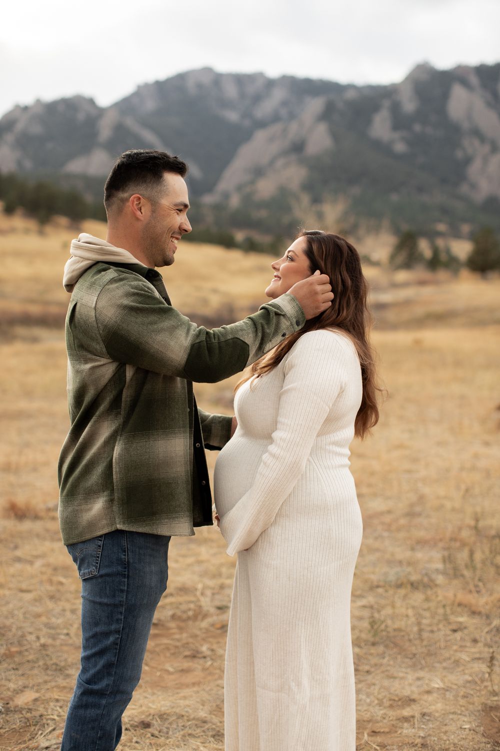 A couple shares a tender moment in a grassy field with dramatic mountains in the background during sunset.