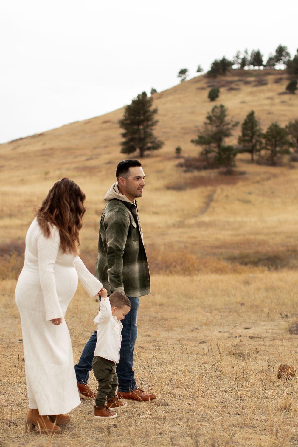 A family in warm clothes standing together on a golden hillside with scattered pine trees in autumn.