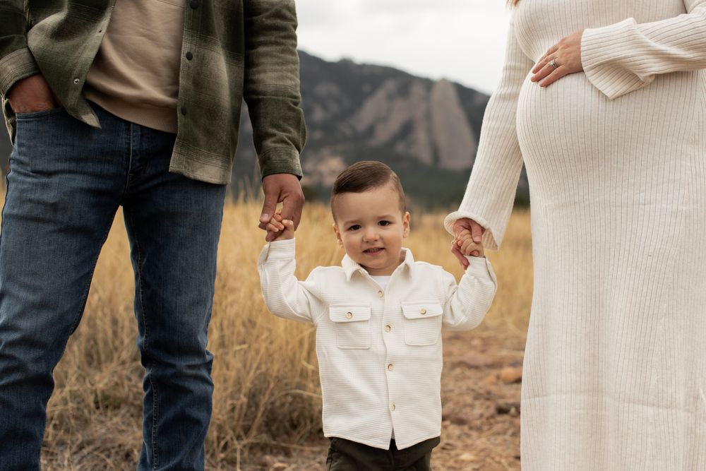 Small child in white shirt holds hands with parents in dry grassland with mountains in background.