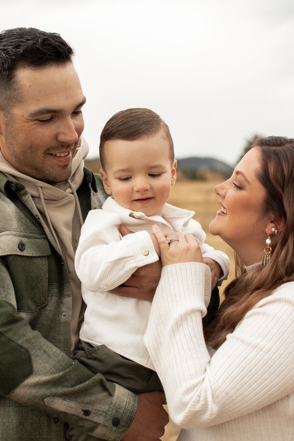 A loving family embraces their baby outdoors while wearing cozy sweaters and casual attire on a cloudy day.