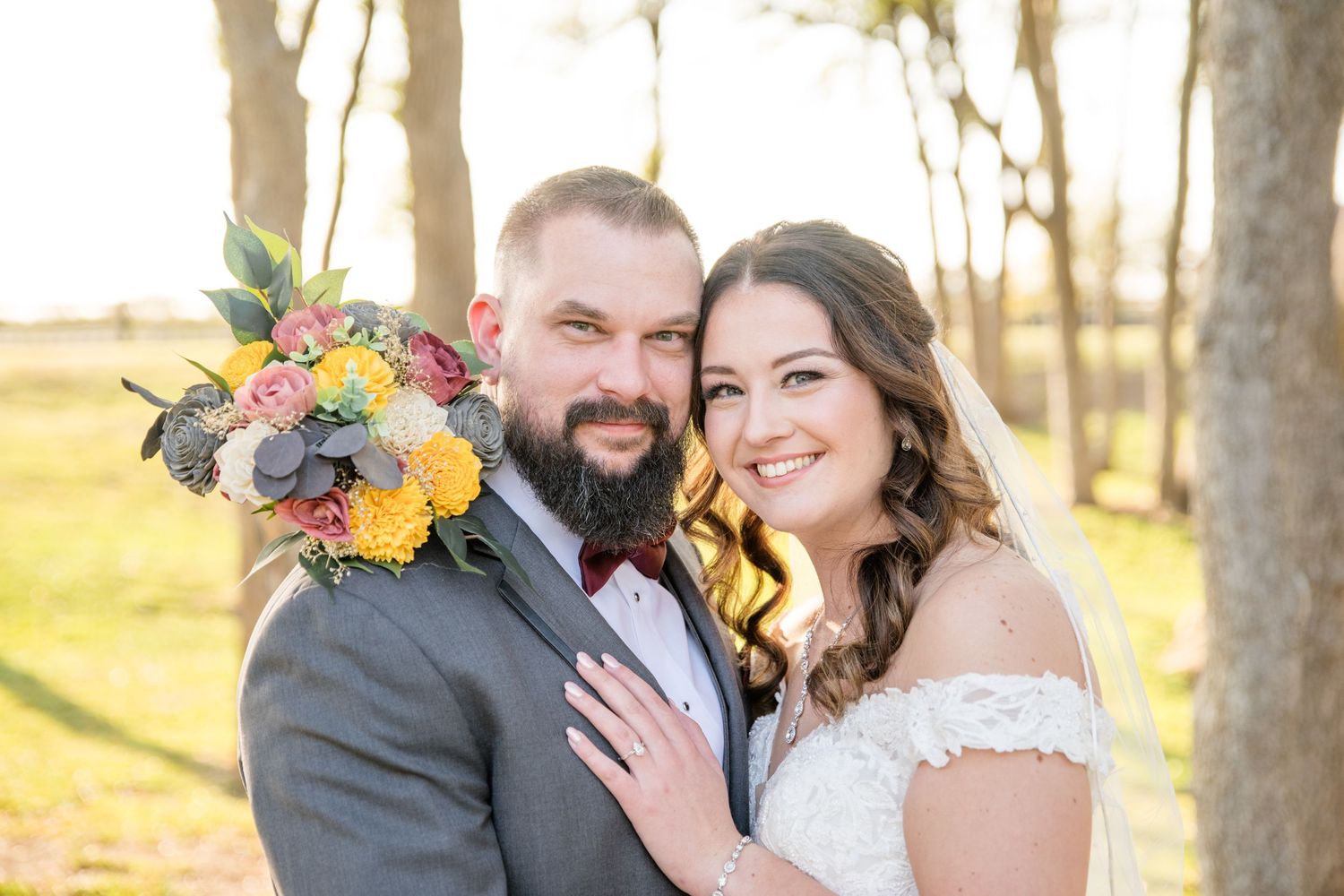 A couple in wedding attire poses outdoors during golden hour with a colorful floral bouquet in their hands.
