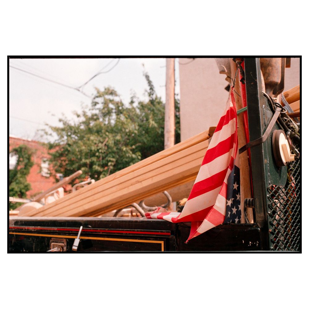 A red and white striped flag hangs from a building with wooden roof beams visible against a pink wall and green trees.