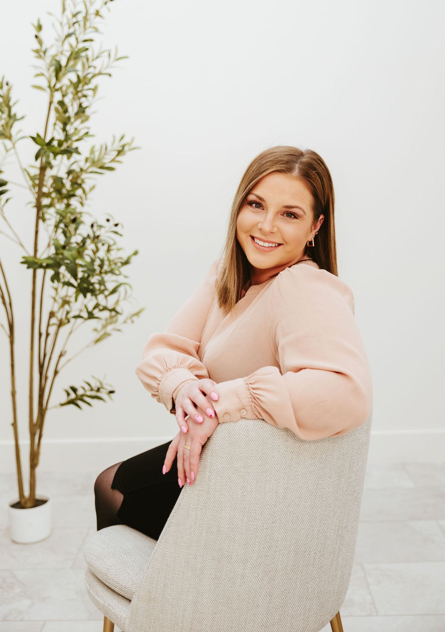 Woman in pink sweater sits elegantly in beige armchair next to potted plant.