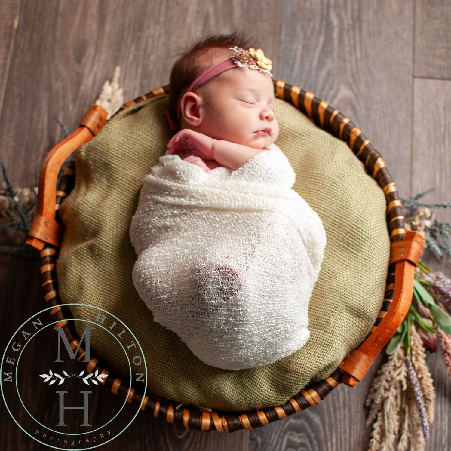 Sleeping baby girl wrapped in white rests in wooden bowl with green cushion and floral decor.