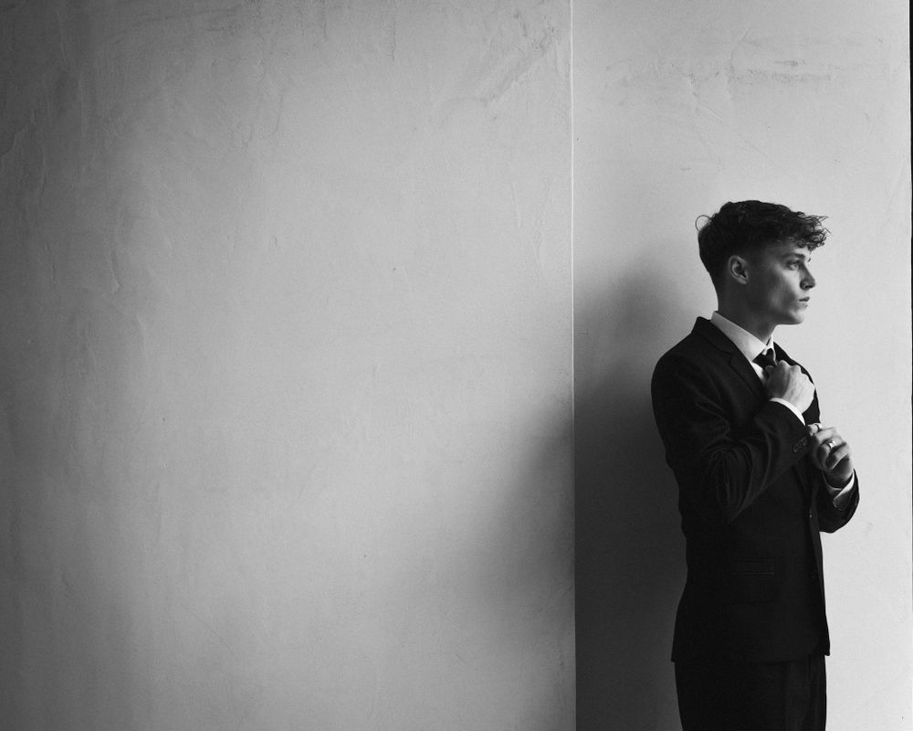 Dramatic black and white portrait of groom getting ready in profile against minimalist white wall.