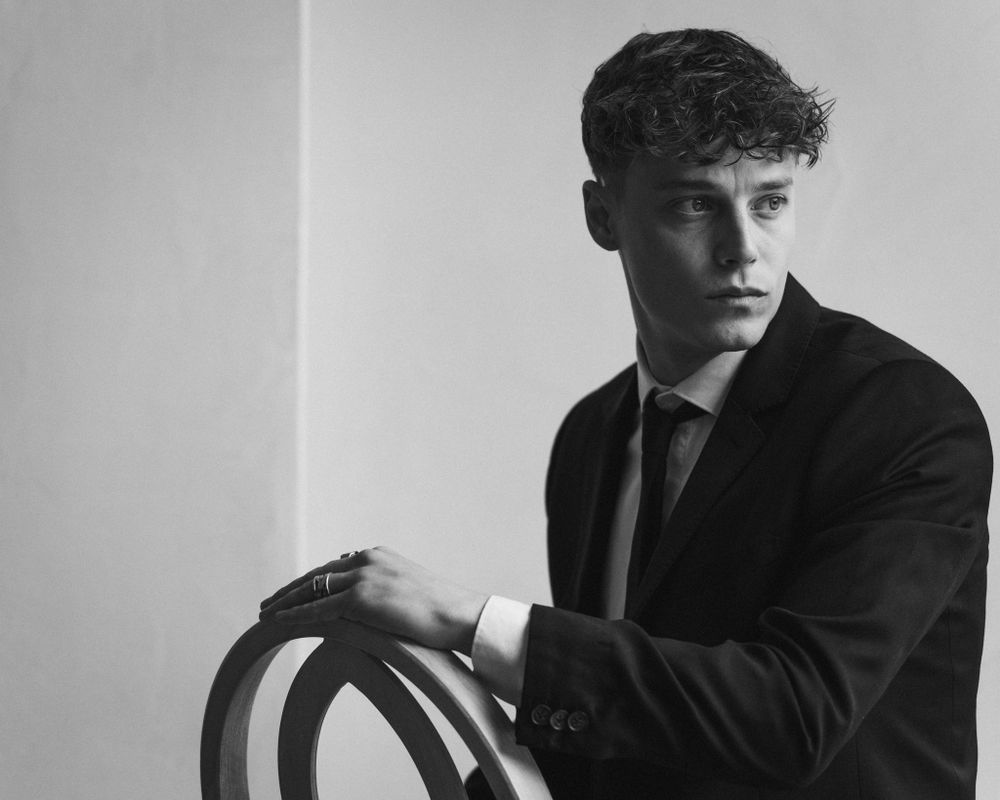 Moody black and white photo of suited figure leaning on curved wooden chair back.