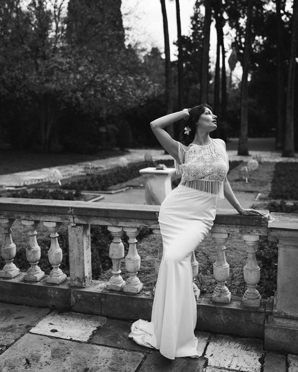 Elegant bride in fitted lace gown poses on stone balustrade in garden setting. Next day wedding photography.