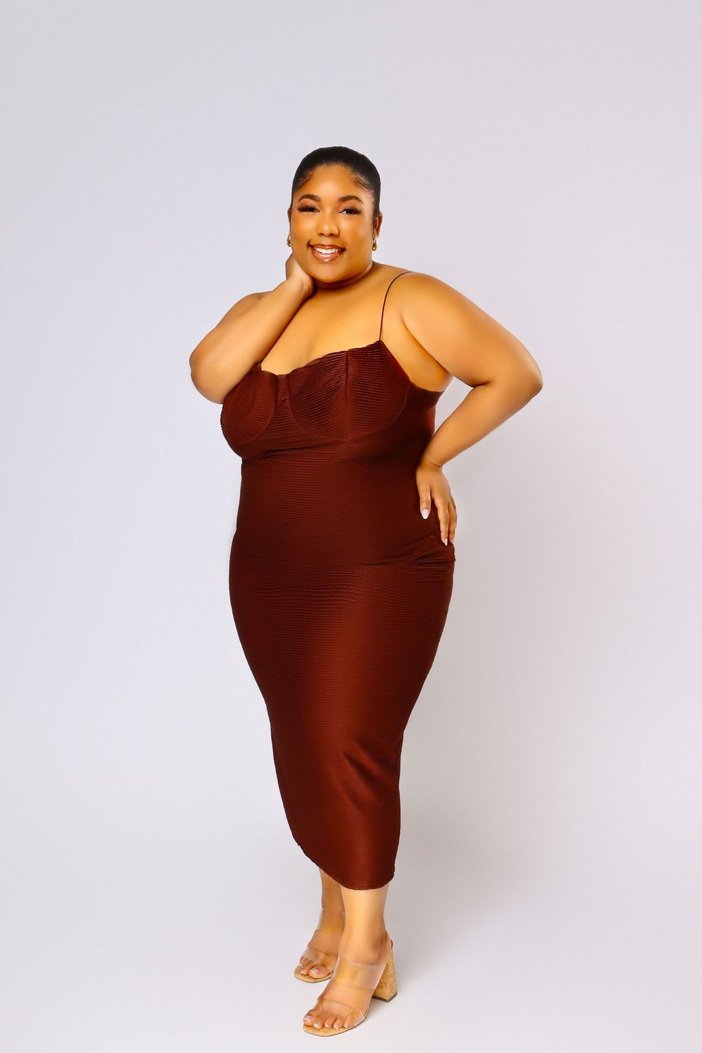 Model in brown fitted dress poses confidently against light background with hand on face.