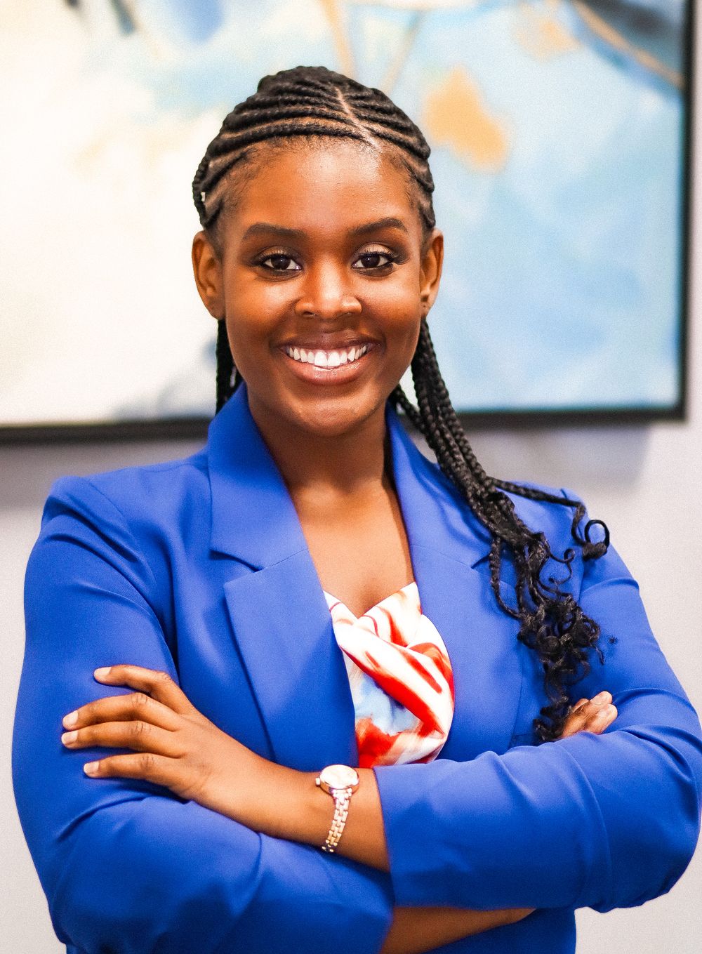 Professional woman in blue blazer and braided hair smiles warmly in office setting.