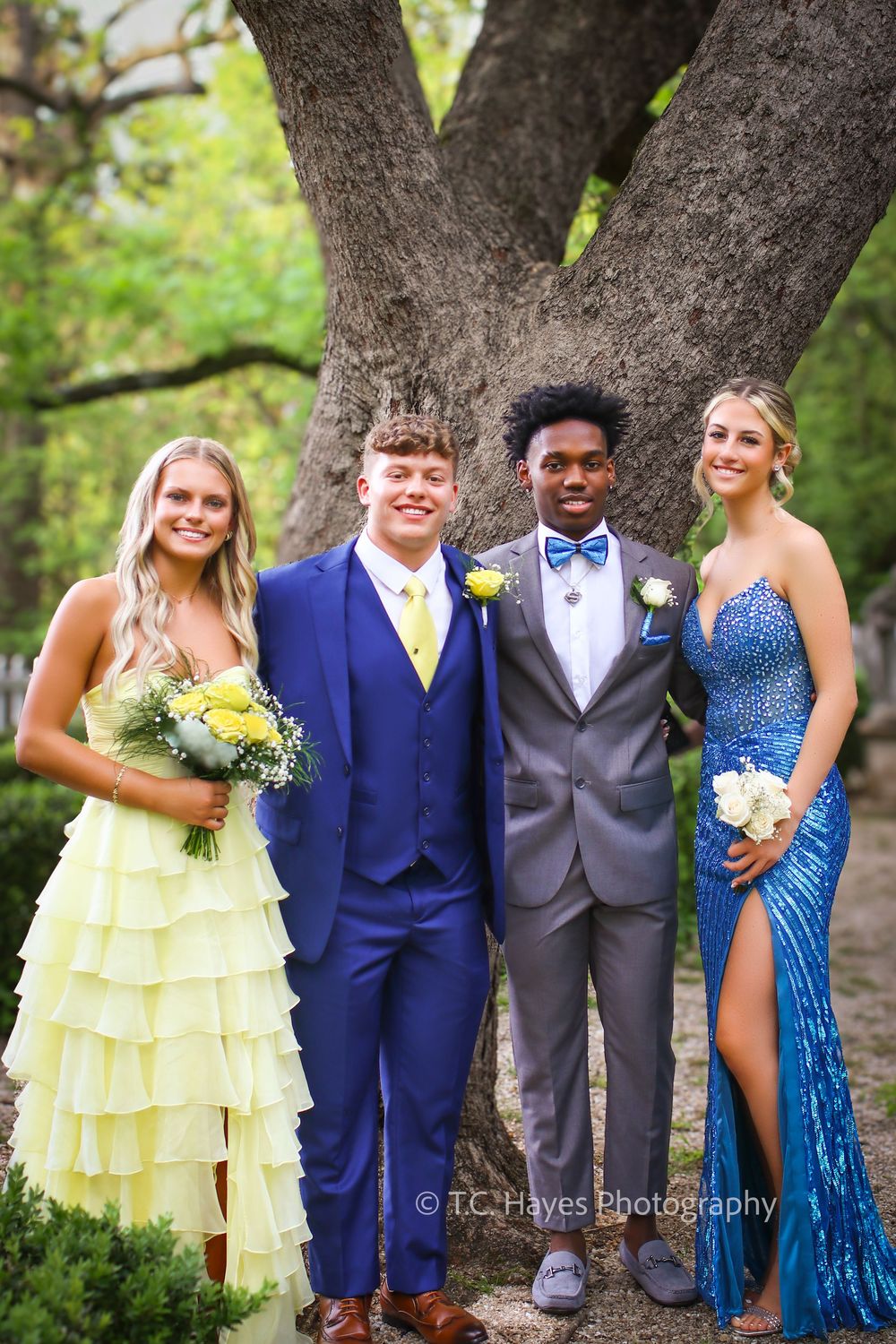 Four students pose in formal attire including yellow and blue dresses and suits at a prom or formal dance event.