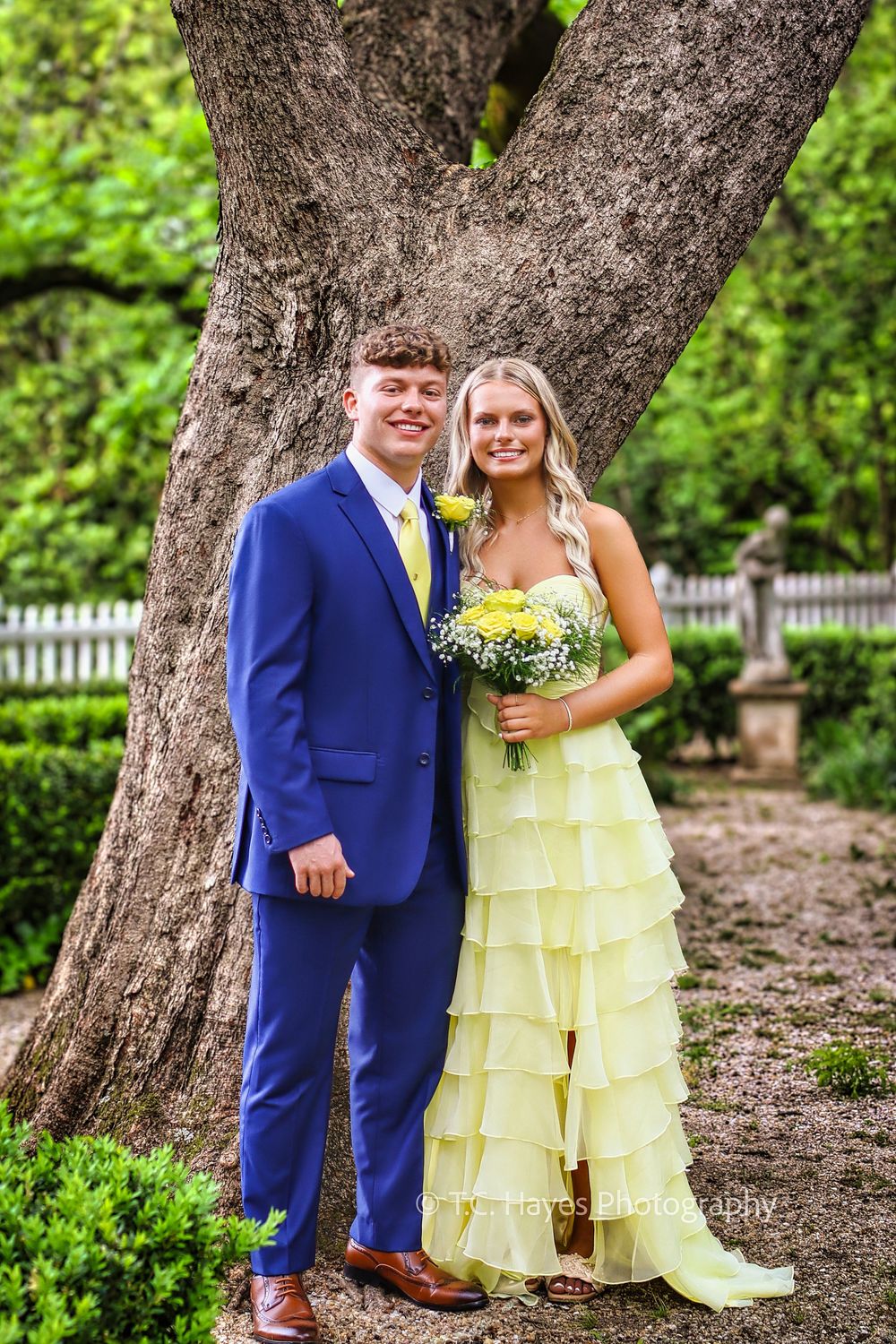 A couple in formal attire poses in front of a large tree trunk, wearing a bright blue suit and yellow tiered gown.