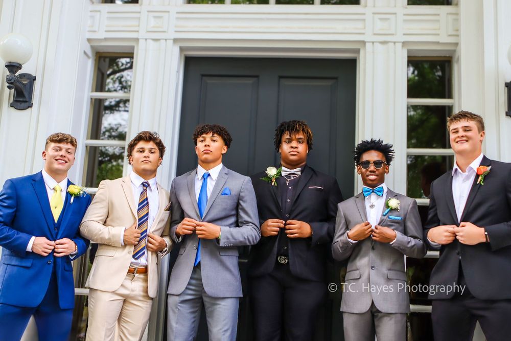 Group of young men in formal suits and ties stand together outside a building entrance for a special occasion.