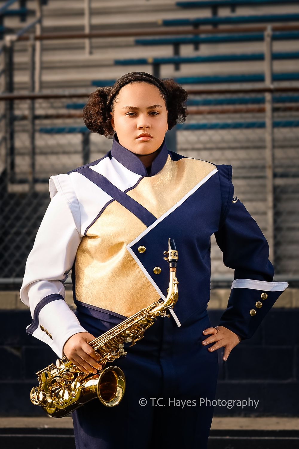 Student wearing a navy and gold marching band uniform stands with saxophone in front of stadium bleachers.