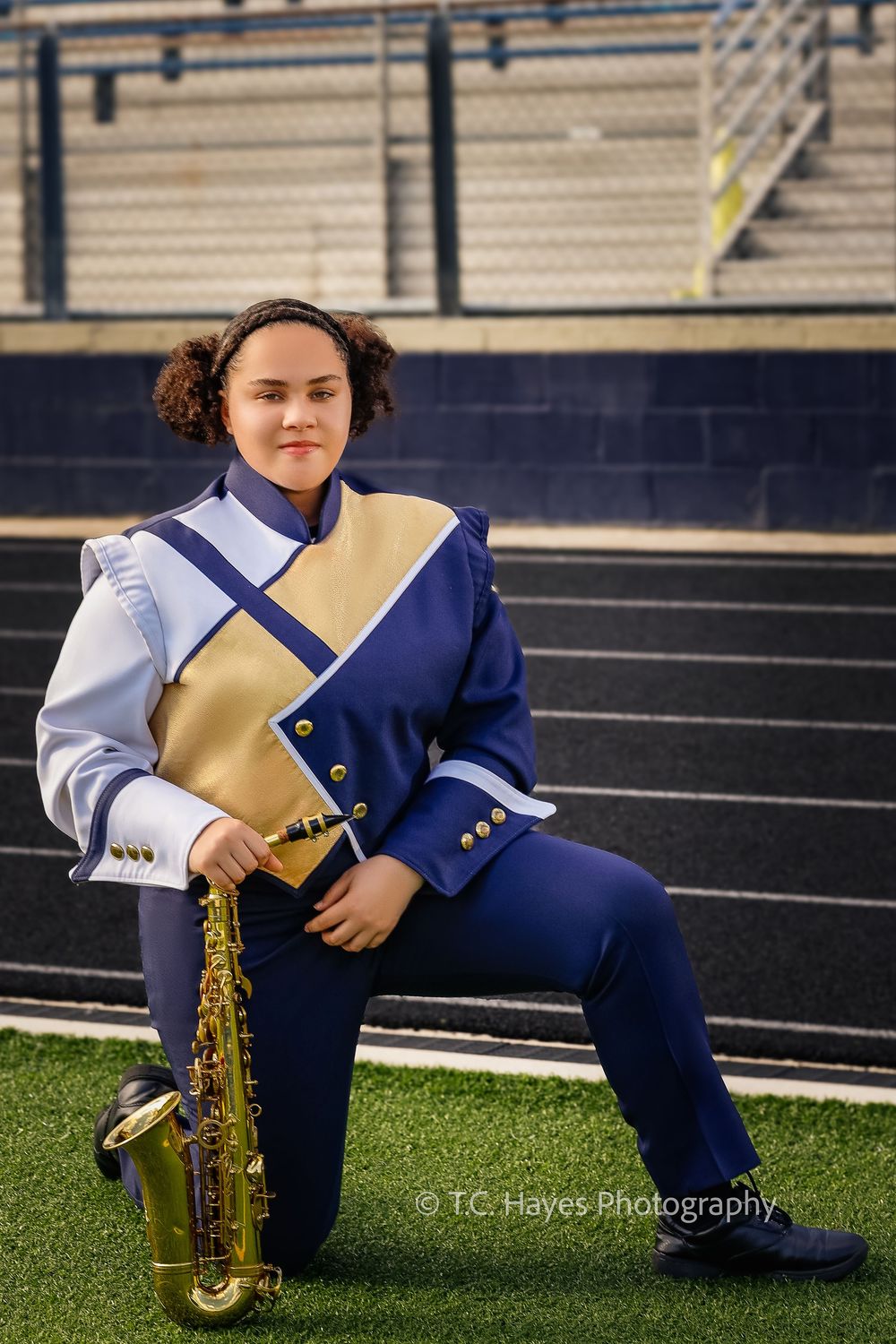 Marching band member in blue and gold uniform poses with saxophone on football field near stadium bleachers.