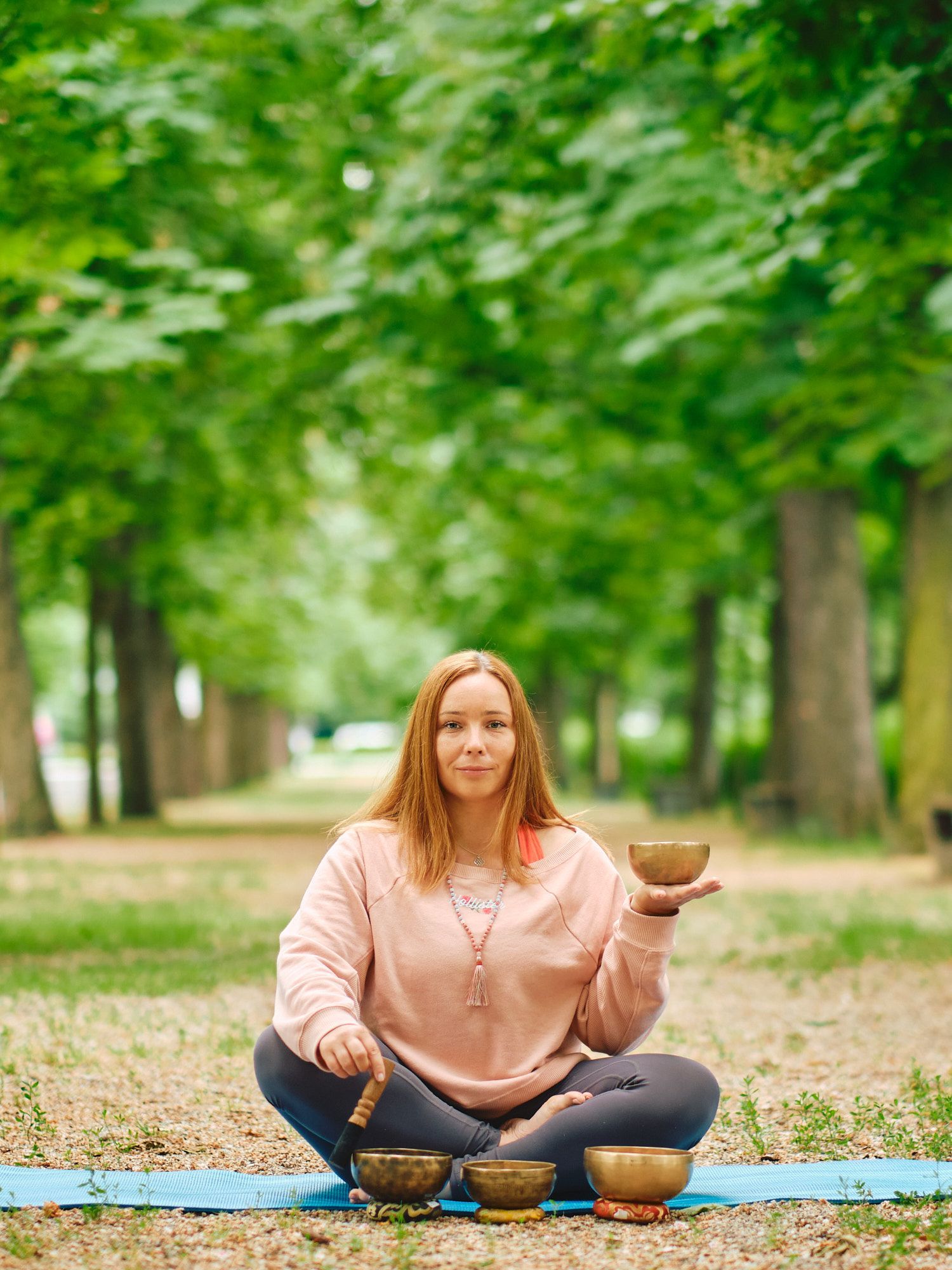 Person in pink sweater sitting cross-legged on yoga mat with singing bowls in park setting surrounded by green trees.
