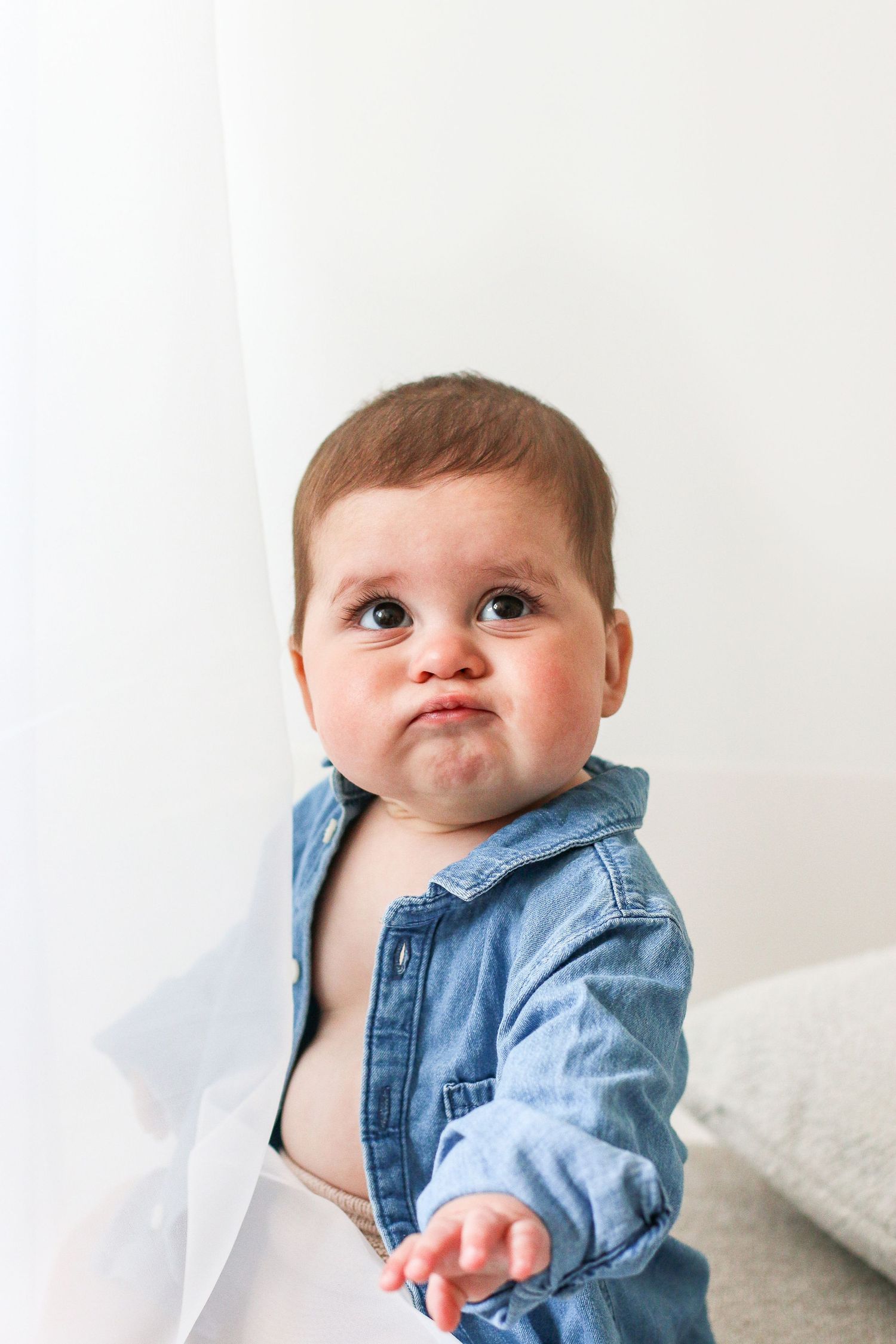 Small baby wearing unbuttoned denim shirt makes adorable chubby-cheeked expression against white background.