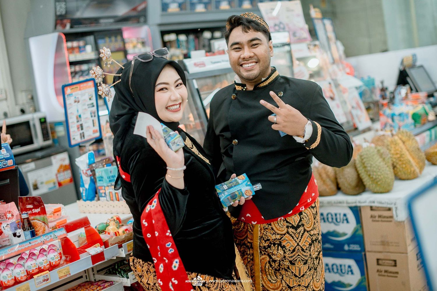 Two cheerful shopkeepers in traditional clothing pose together at their grocery store showing peace signs.