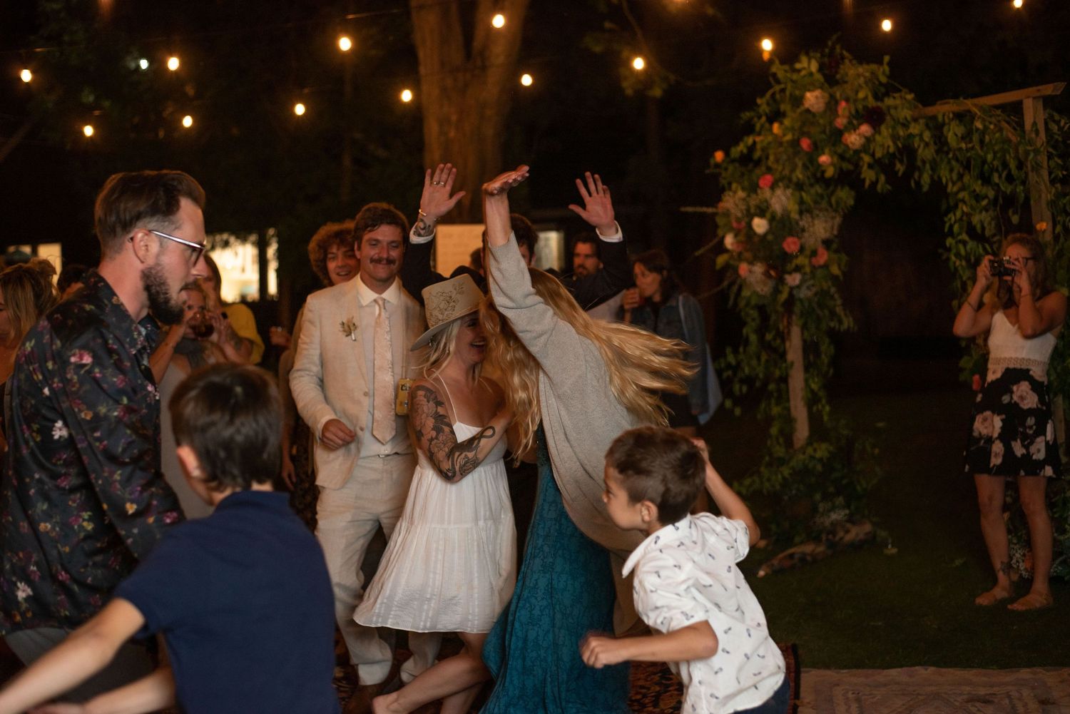 Wedding guests dance and celebrate at an outdoor evening reception with string lights and greenery decorations.