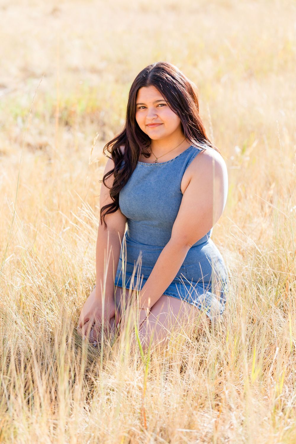 Young woman in blue dress sitting in golden field of tall dry grass, smiling softly in warm sunlight.