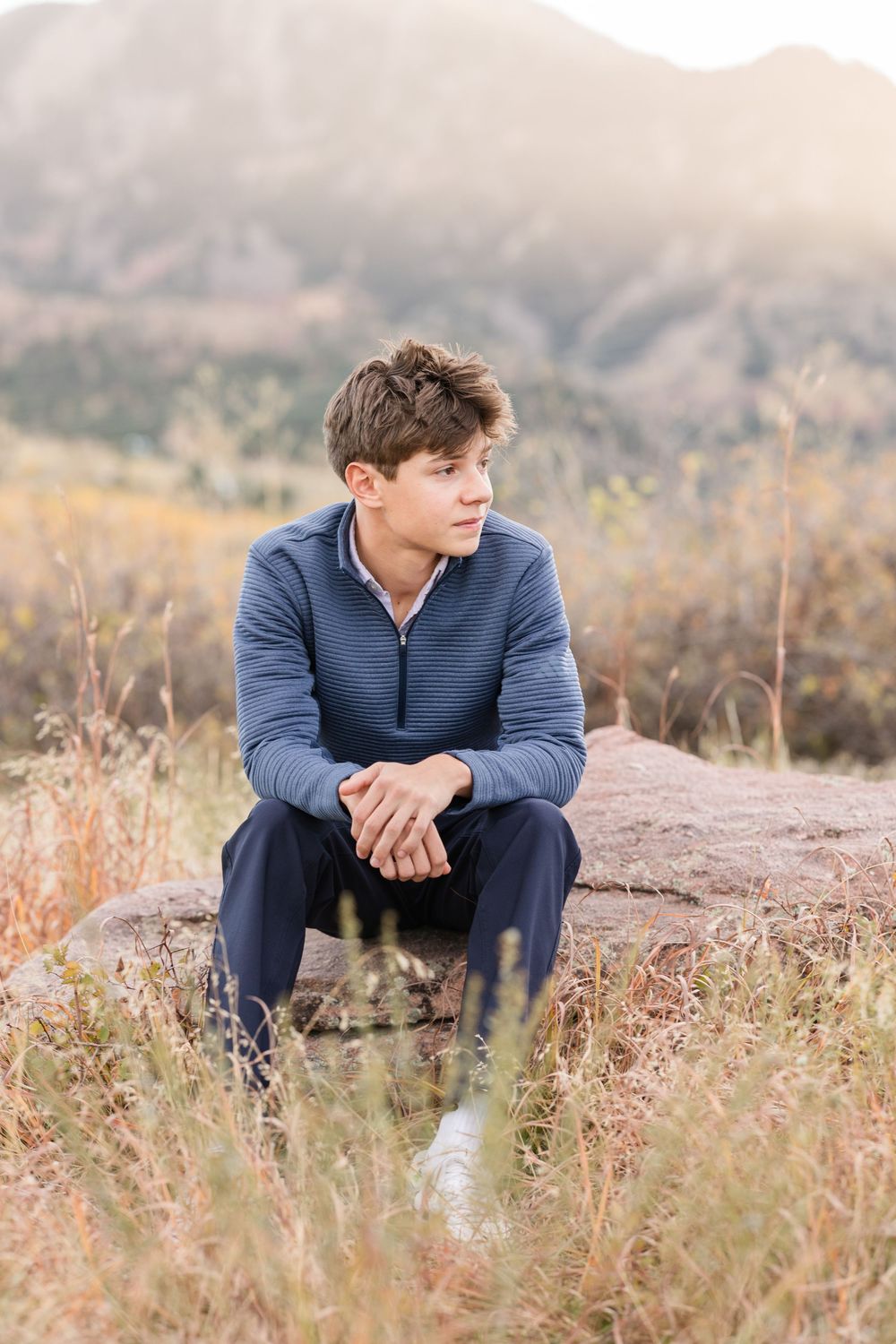 Person in blue sweater sitting thoughtfully in autumn field with golden grass and mountain backdrop.