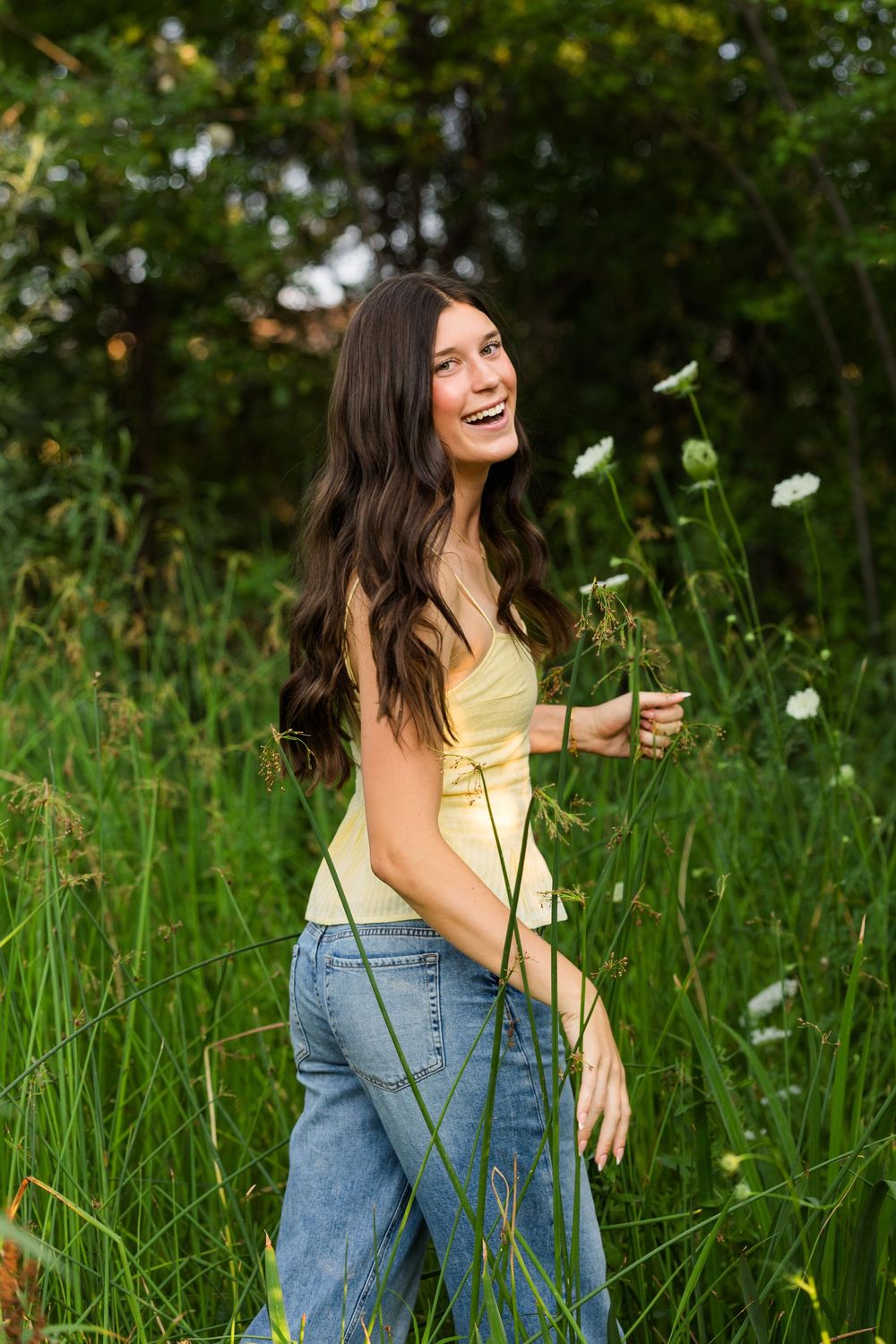 Young woman with long dark hair laughs in a lush green field, wearing a yellow top and jeans, holding wildflowers.