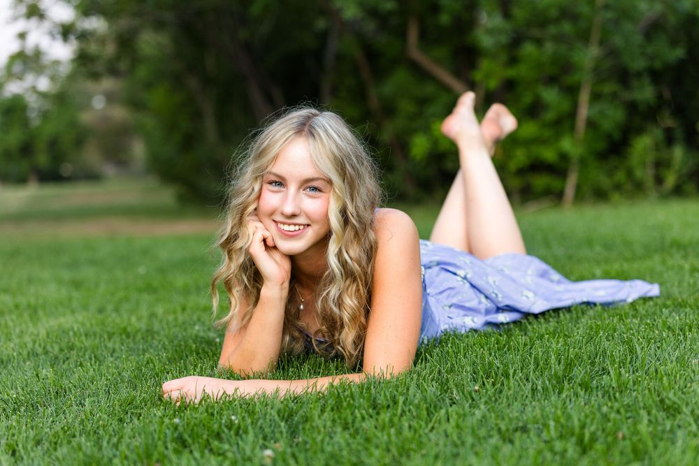 Young blonde woman in blue dress lying on green grass, smiling cheerfully in a sunny outdoor park setting.