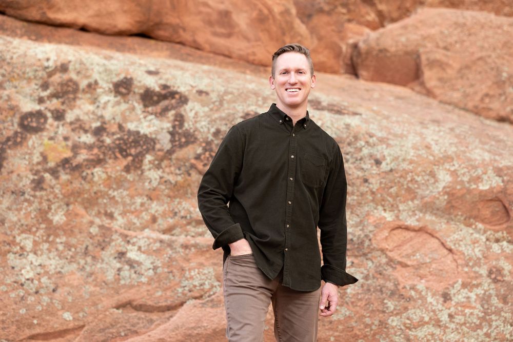 Someone in a black button-down shirt and gray pants poses outdoors against red rock formations in natural lighting.