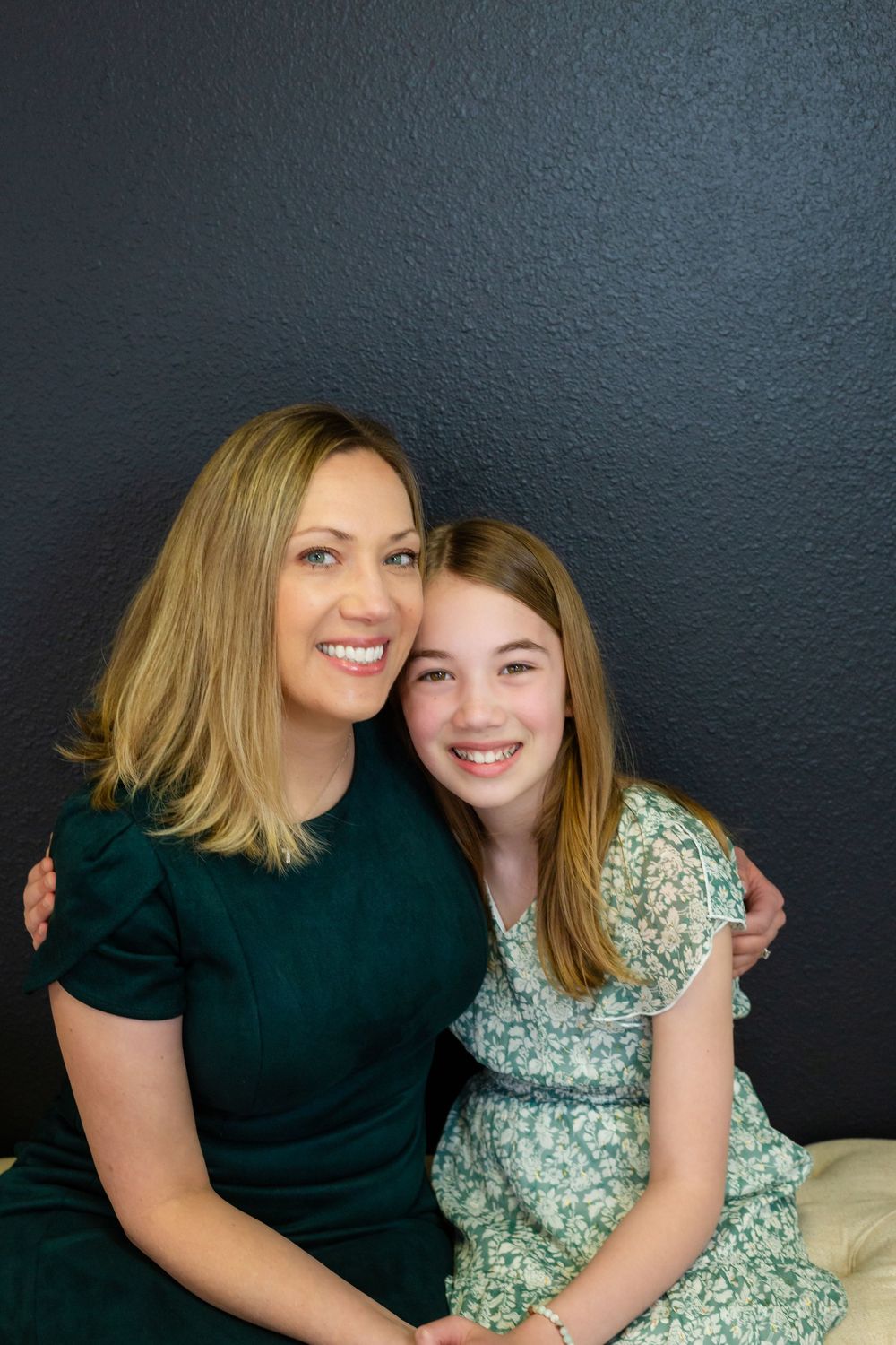 Two people in contrasting outfits of green and floral print sit close together against a dark wall, sharing warm smiles.