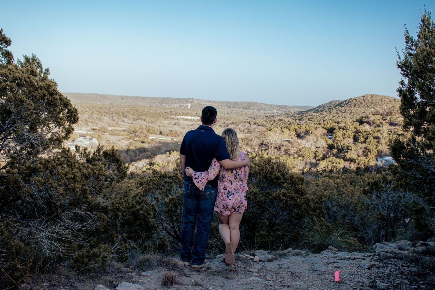 Two people stand together on a hilltop overlook, taking in sweeping views of an arid desert landscape below.