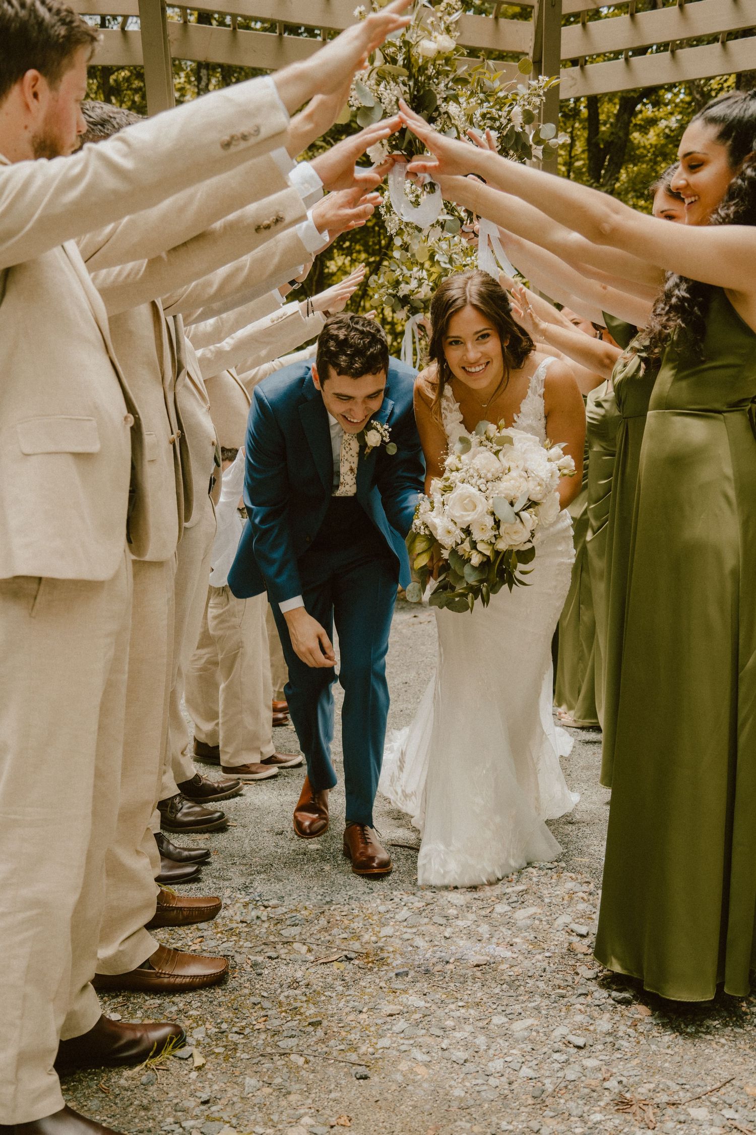 Wedding party forms celebratory arch tunnel while newlyweds duck underneath with bouquet in hand.