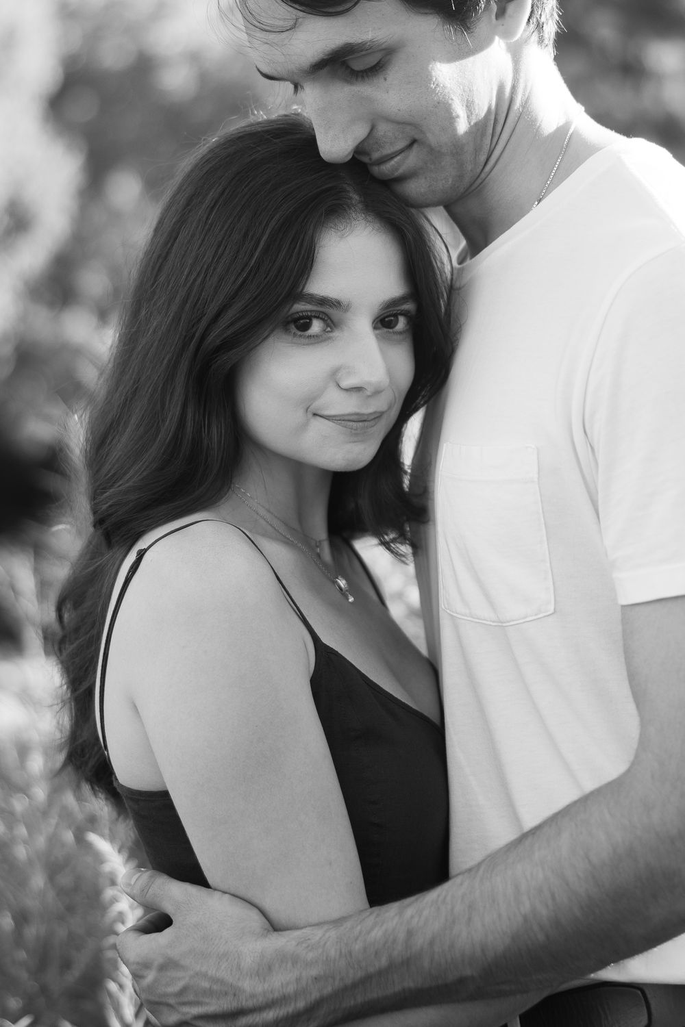 A romantic black and white portrait of a couple embracing outdoors in soft natural lighting.
