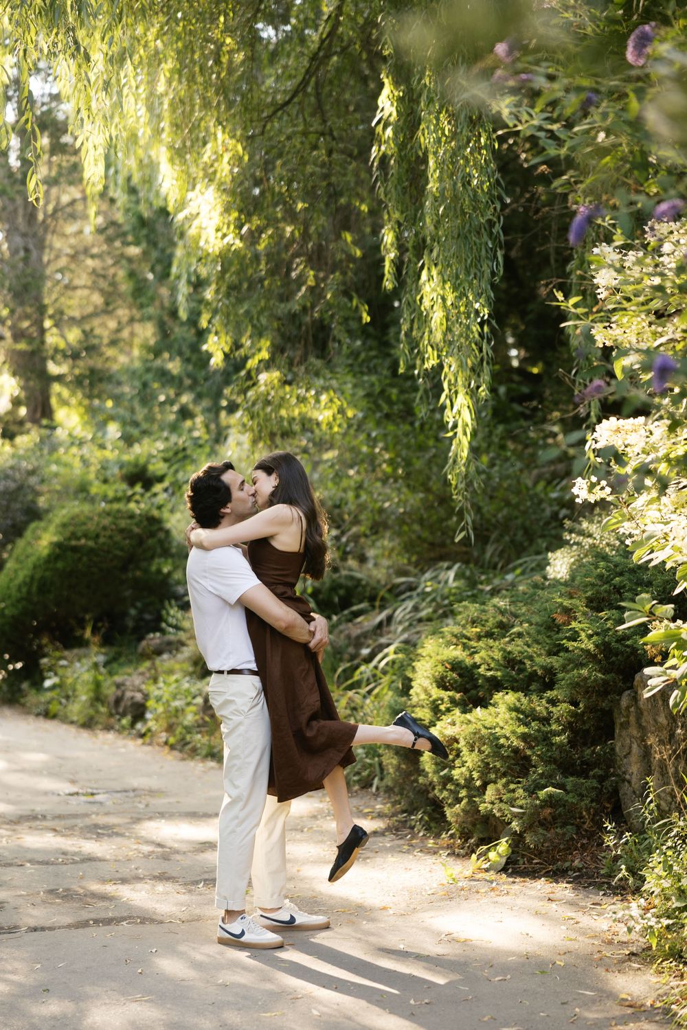 A romantic couple embraces and spins joyfully on a sunlit garden path surrounded by lush greenery and willow trees.