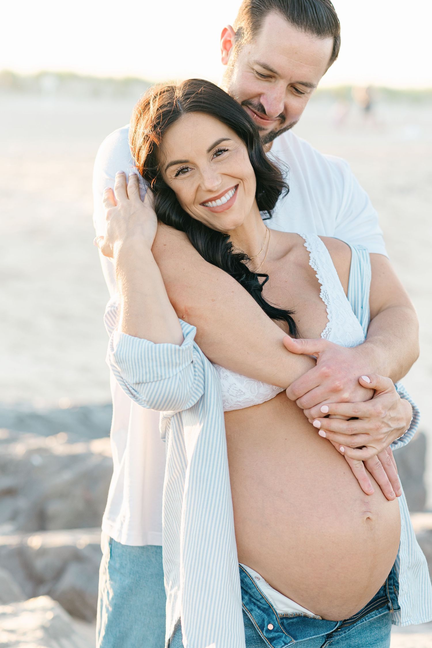 Pregnant woman in baggy jeans and oversized button down shirt cuddles with husband during a beach maternity session in Monmouth County, NJ.