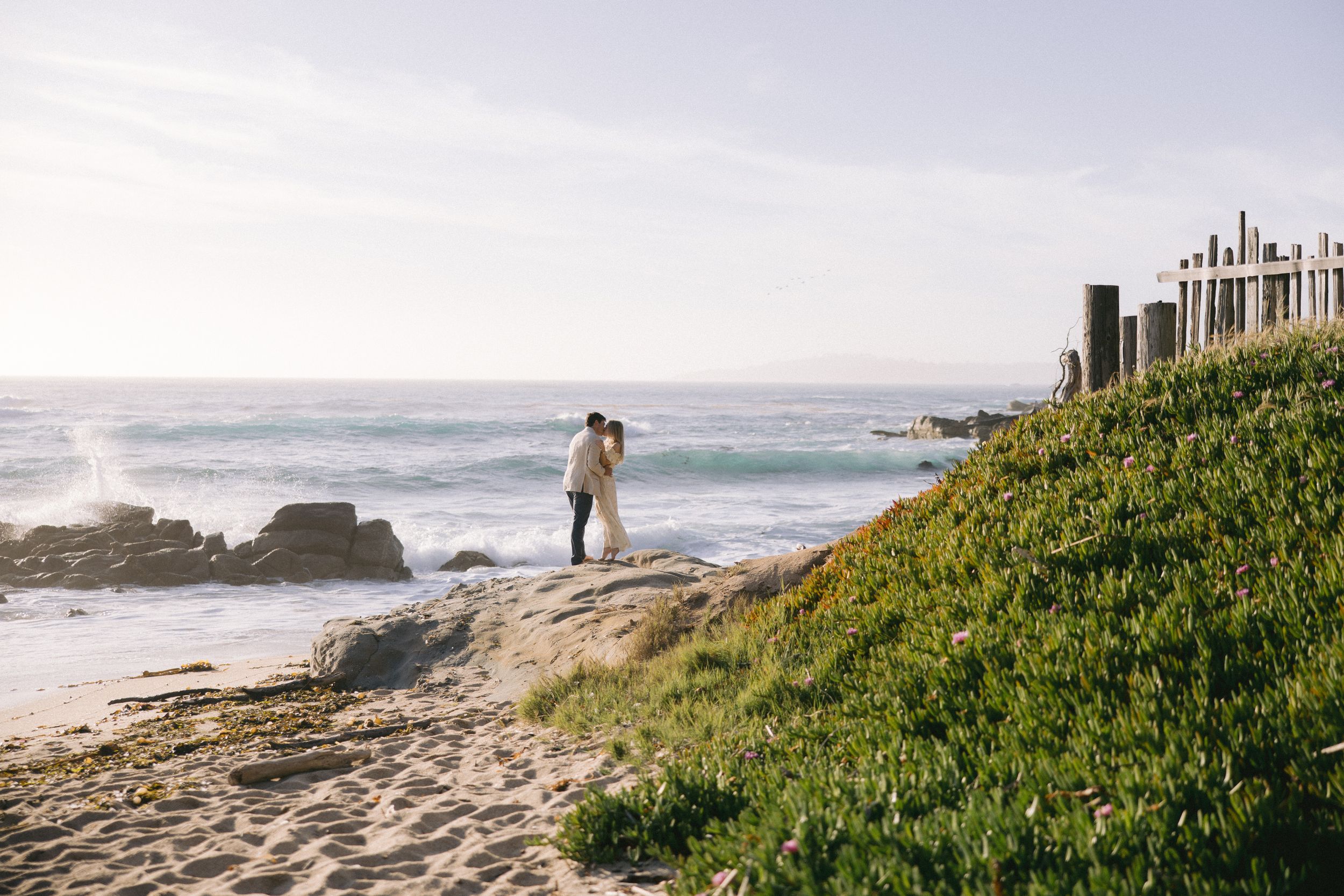 A coastal scene with someone standing on rocky shoreline overlooking the Pacific Ocean with green grass and houses nearby.