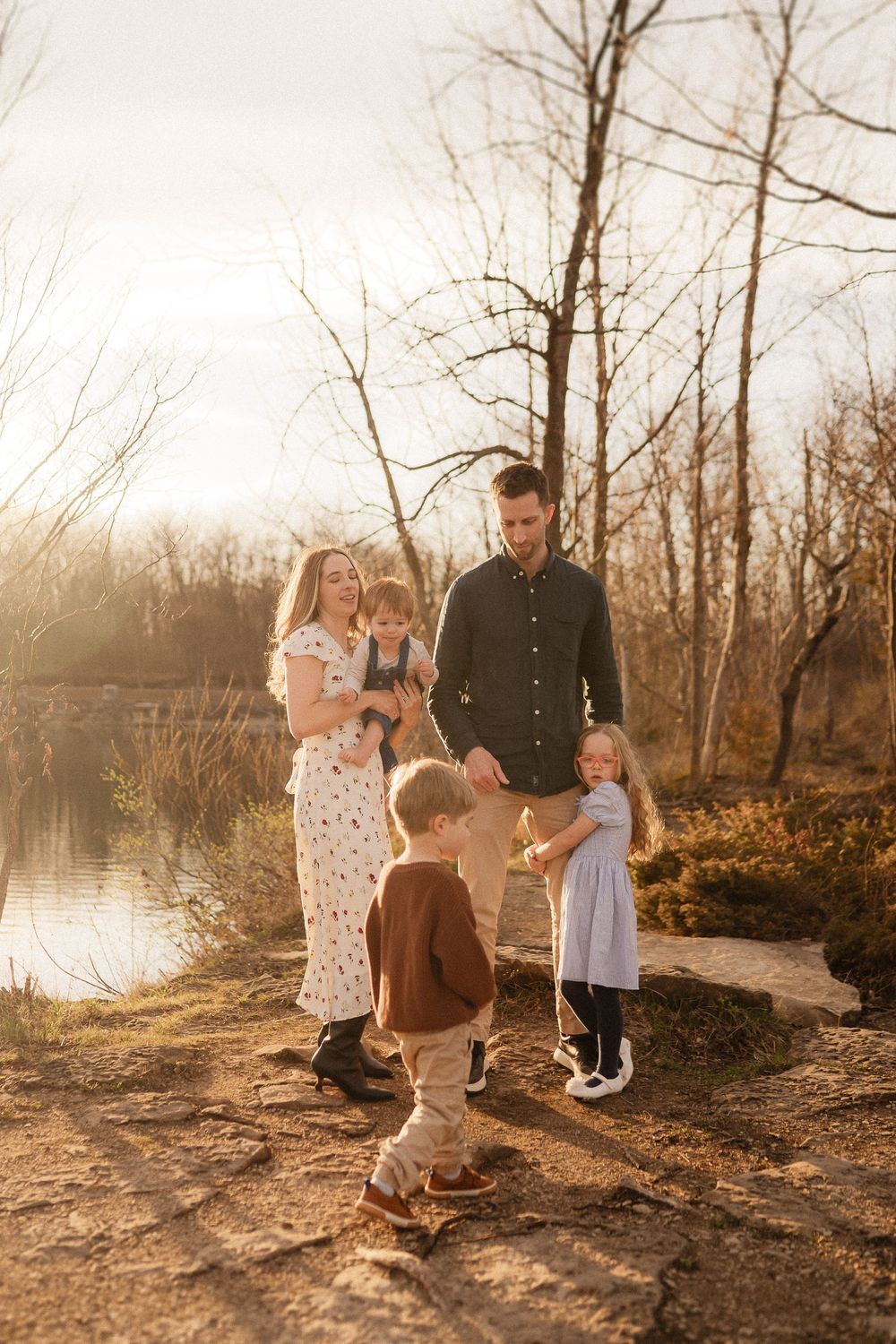 Family gathered on a sunlit path by a lake during golden hour with bare trees in the background.